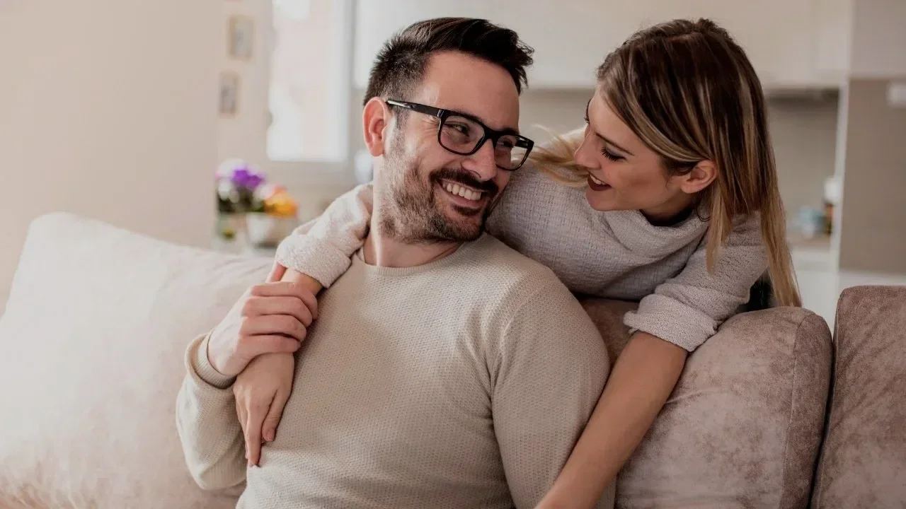 A smiling man with glasses and a woman sitting closely on a beige couch, sharing a joyful moment indoors.