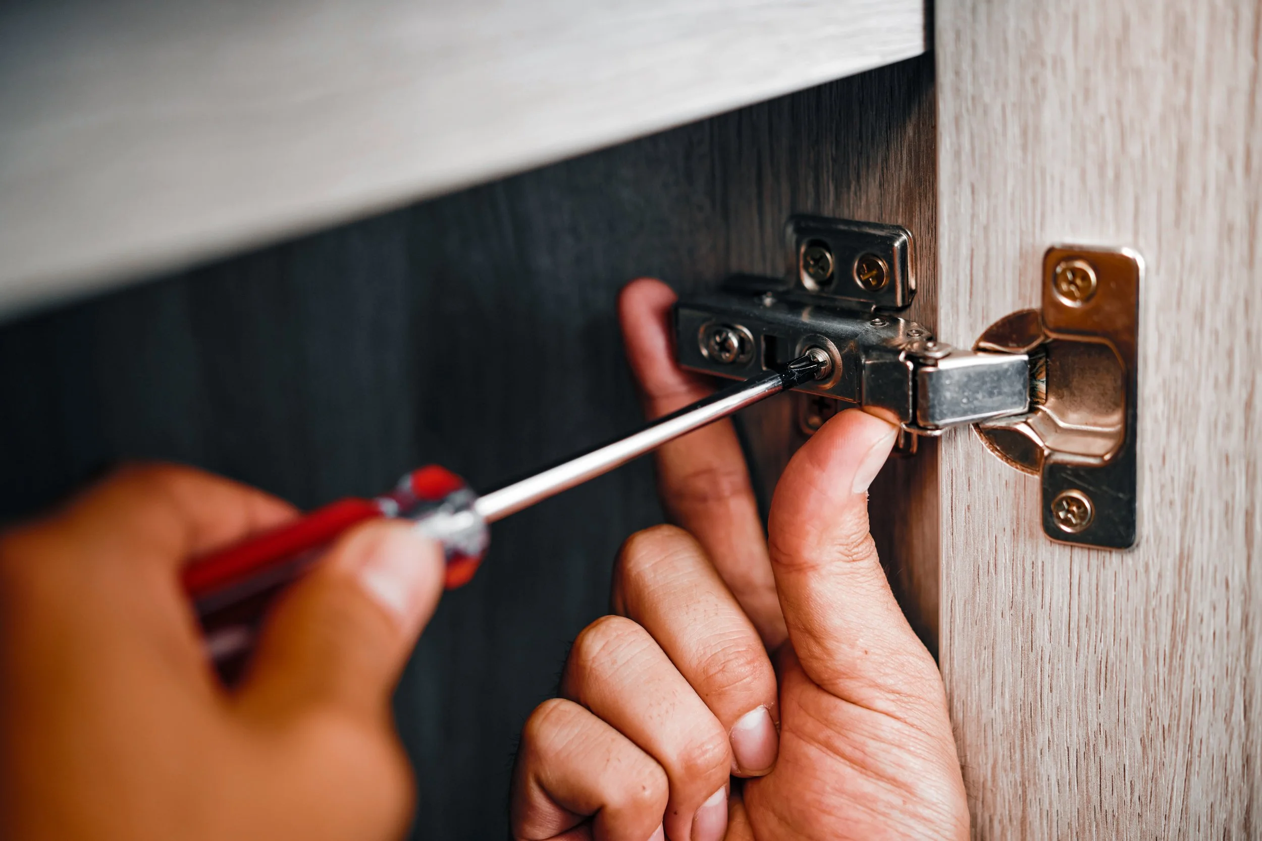 Person using a screwdriver to adjust a cabinet hinge inside a wooden cabinet.