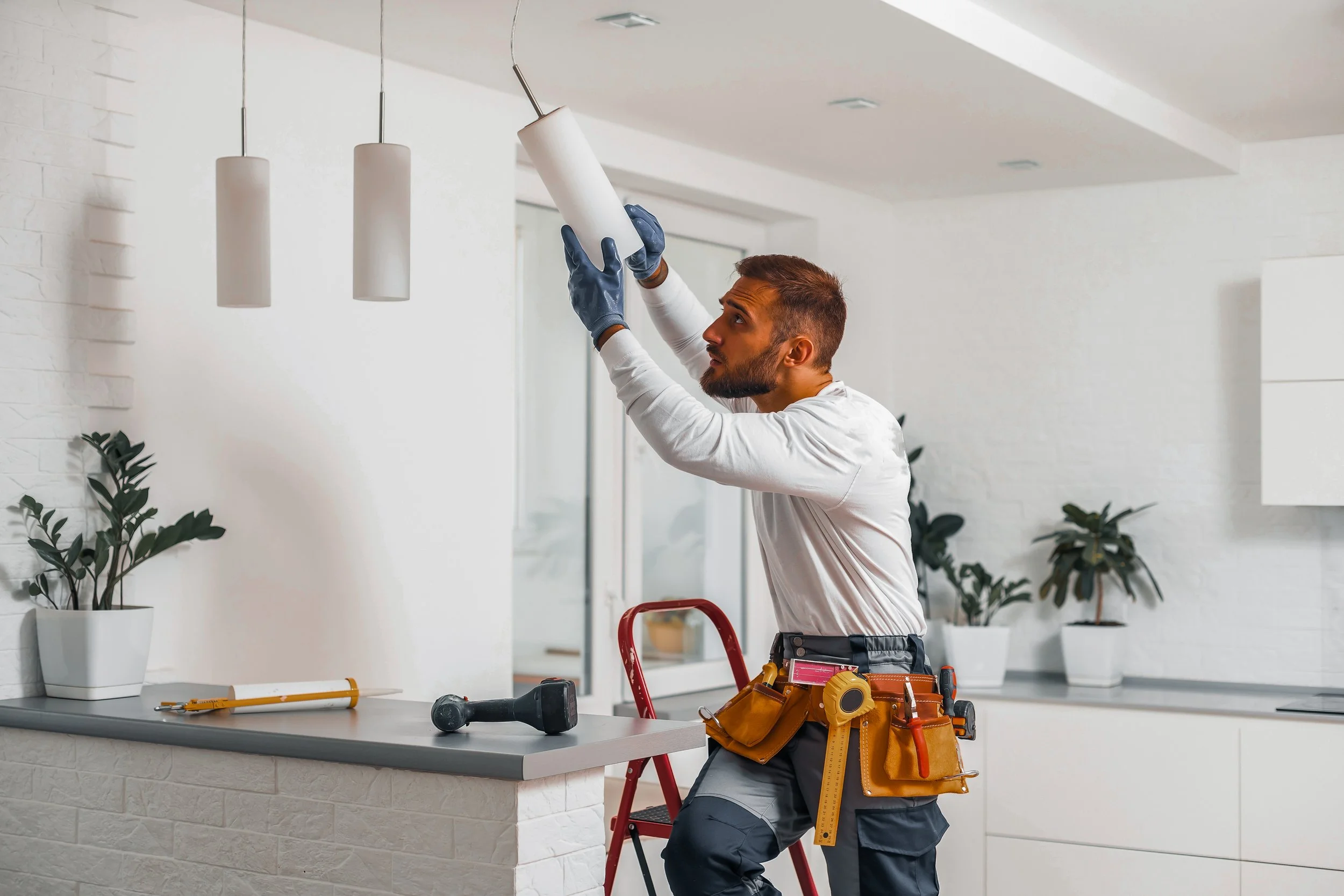 Electrician installing a pendant light in a modern kitchen, using a step ladder and wearing a tool belt with various tools.