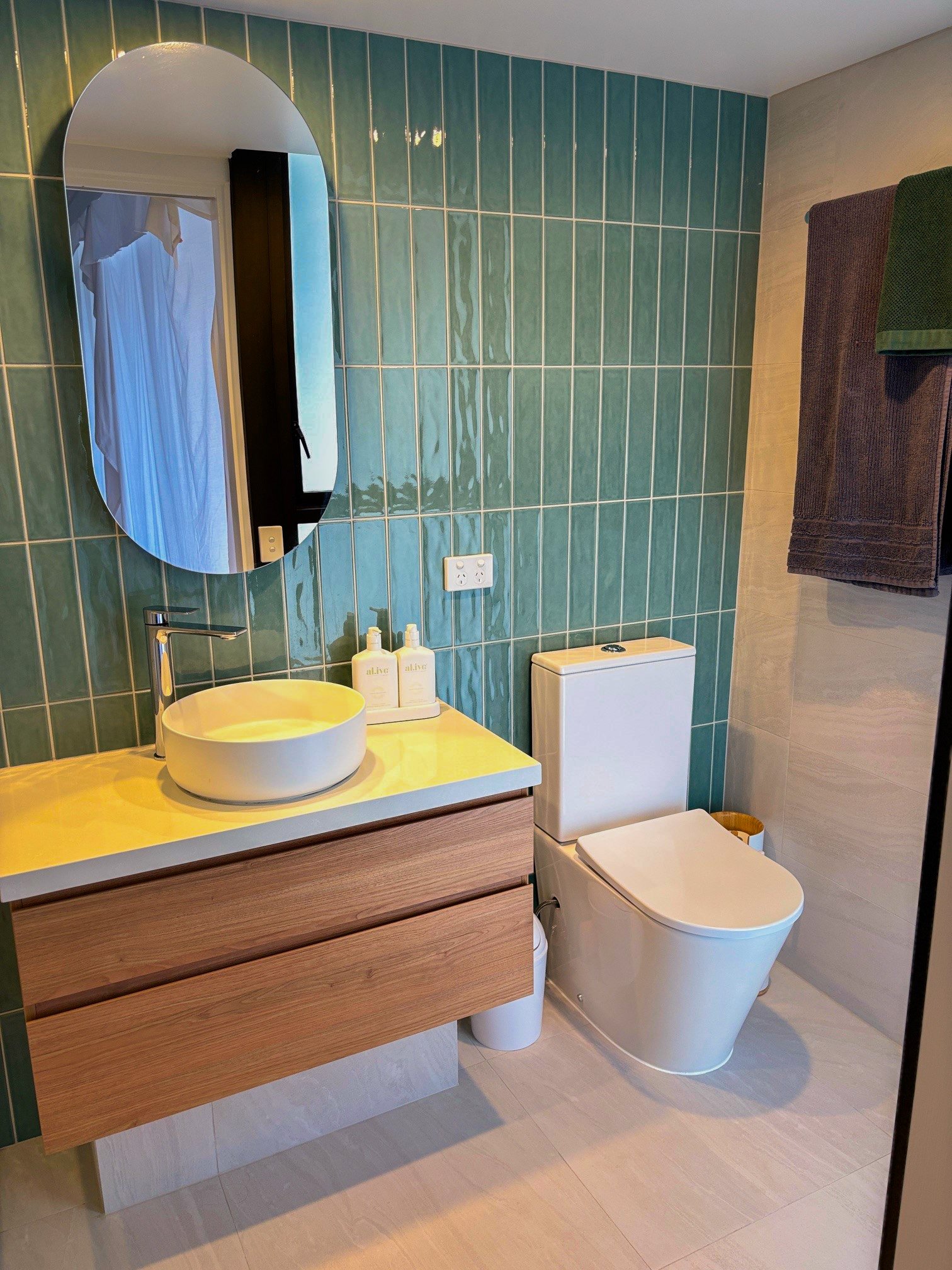 Modern bathroom with a wall-mounted wooden vanity featuring a round vessel sink and a curved mirror. The room has teal tiled walls, a white floor, and a white toilet. Towels hang on a rack, and toiletries are on the vanity.