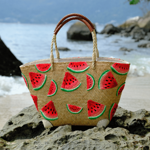 Front view of the Phatsajira Watermelon Bites tote placed on sunlit beach rocks, showcasing its hand-embroidered watermelon design against natural Krajood weave.