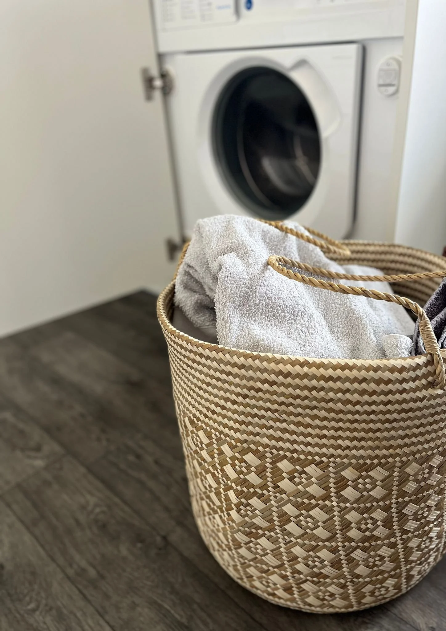 The Sailom basket, filled with grey laundry, is placed in front of a modern white washing machine.
