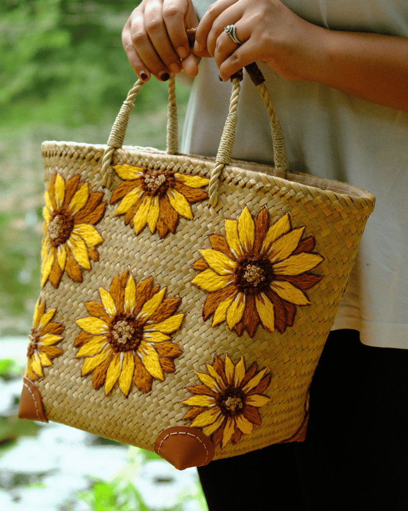Sunflower-embroidered straw tote with leather-wrapped handles shown in an outdoor tropical setting, reflecting the bag’s summer inspiration and artisanal charm.