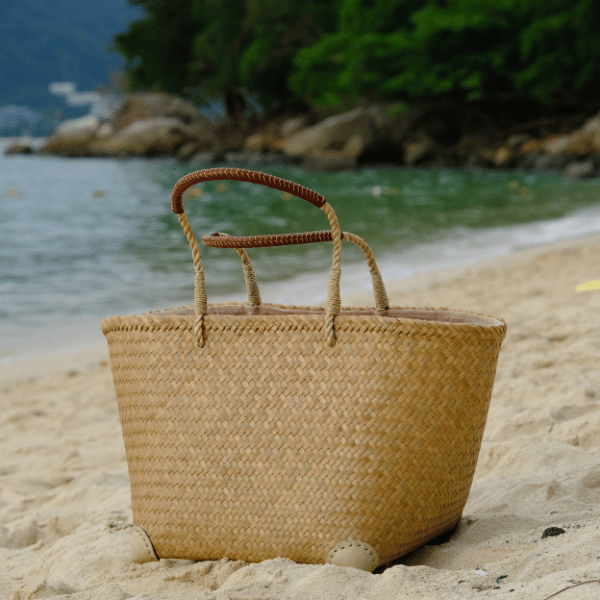 Rewadee tote positioned upright on white sand beside the sea, framed by natural scenery for a timeless coastal feel.