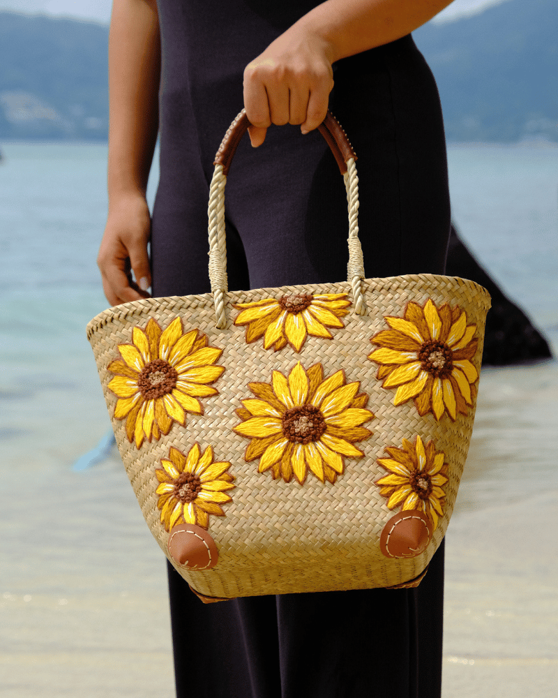 Woman standing by the beach holding the Phatsajira Sunflower tote bag, highlighting its structured shape and vibrant floral embroidery against a coastal backdrop.