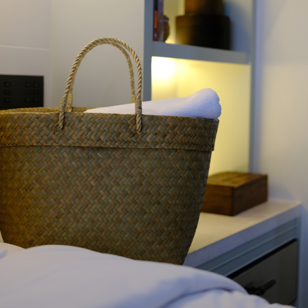 Tung Yai basket positioned on a bedside table, softly lit by a nearby lamp, with folded towels neatly inside.