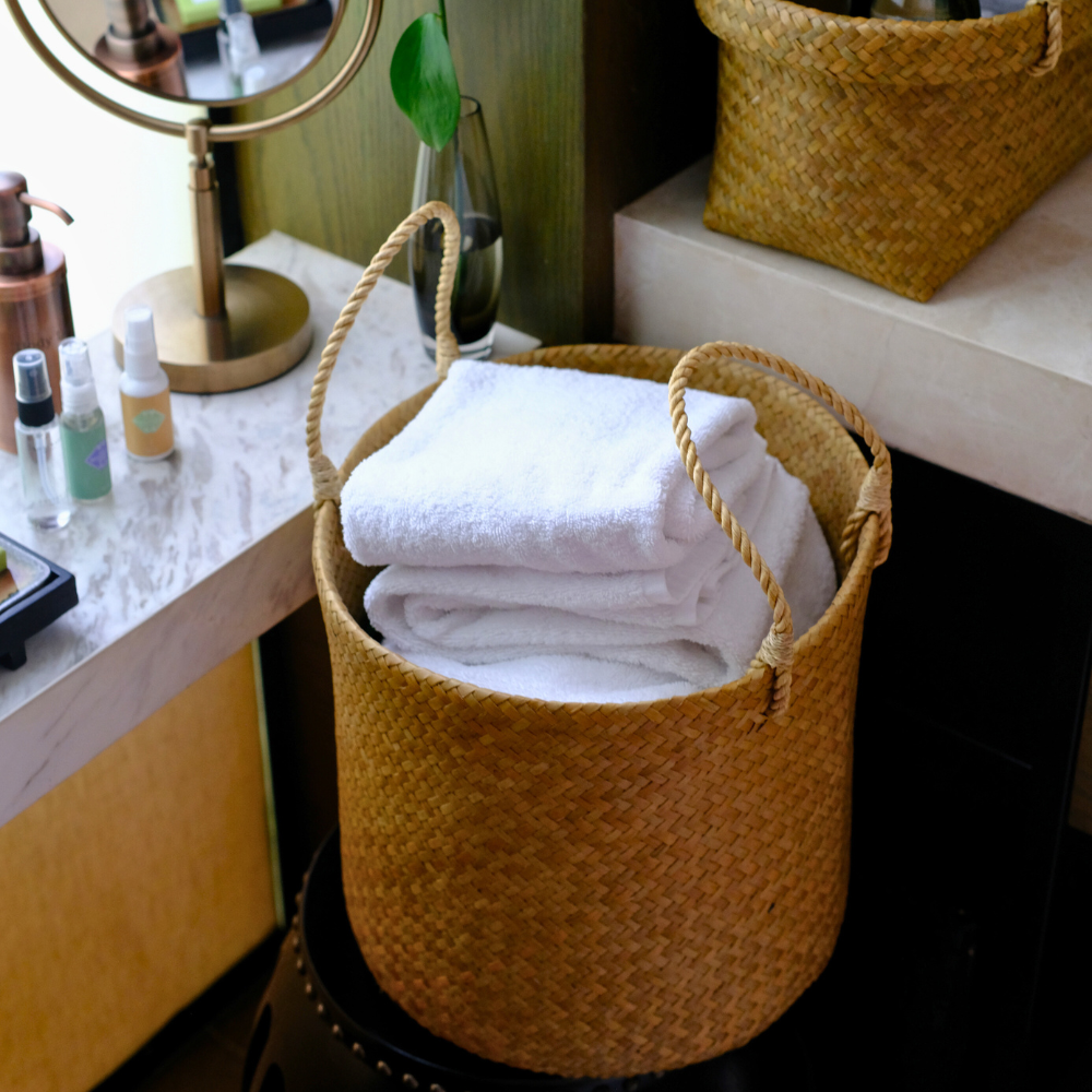 Close-up of the Tung Pha basket beside a bathroom sink with folded towels inside, surrounded by daily-use items like a mirror and skincare products.