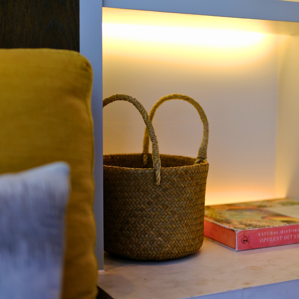 The Tung Dek basket styled on a bedroom shelf beside a stack of books, softly illuminated to show its compact form and fine woven texture.