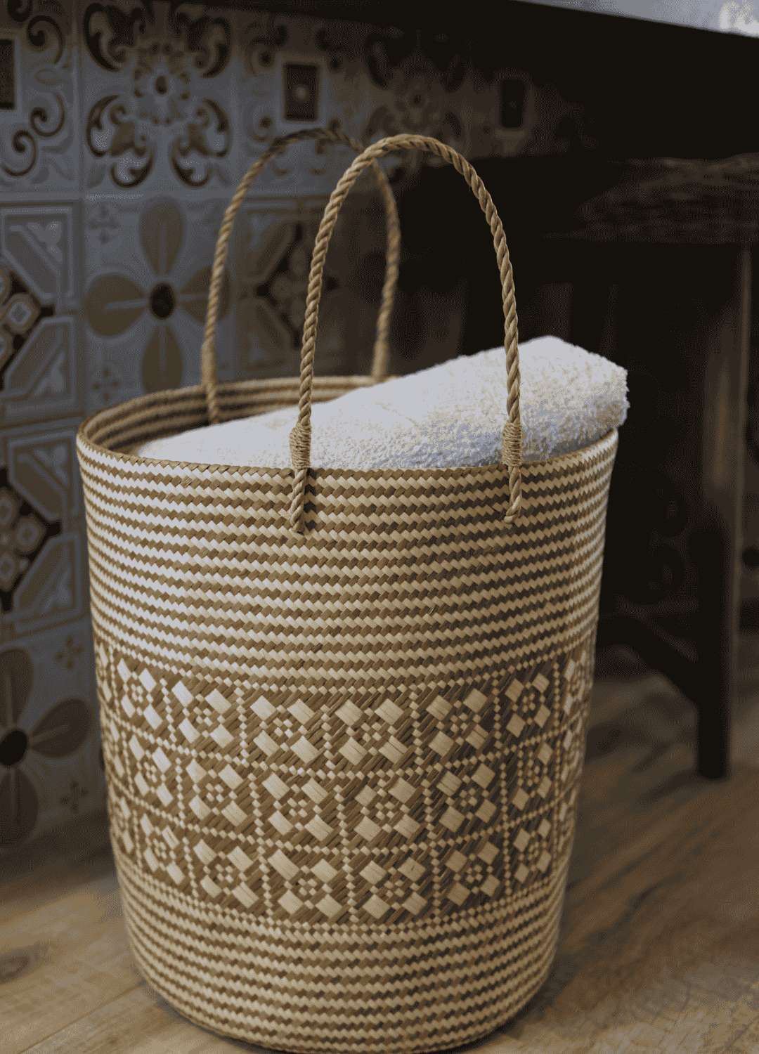 A tall, handwoven laundry basket with geometric patterns sits neatly against a patterned tile wall, filled with a white towel.