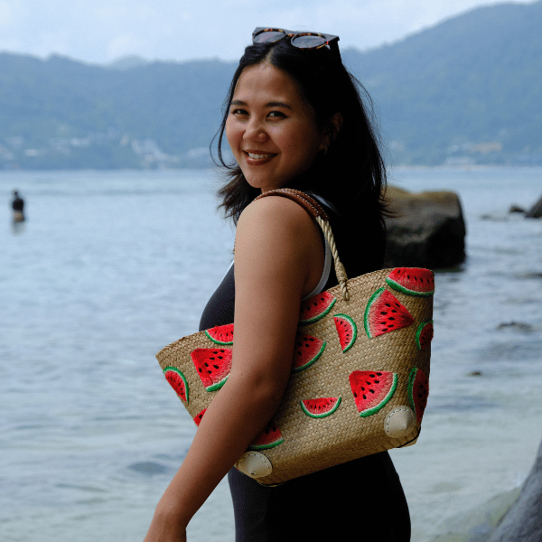 Woman smiling as she wears the Watermelon Bites tote on her shoulder by the sea, showing its scale and comfortable leather-wrapped handles.