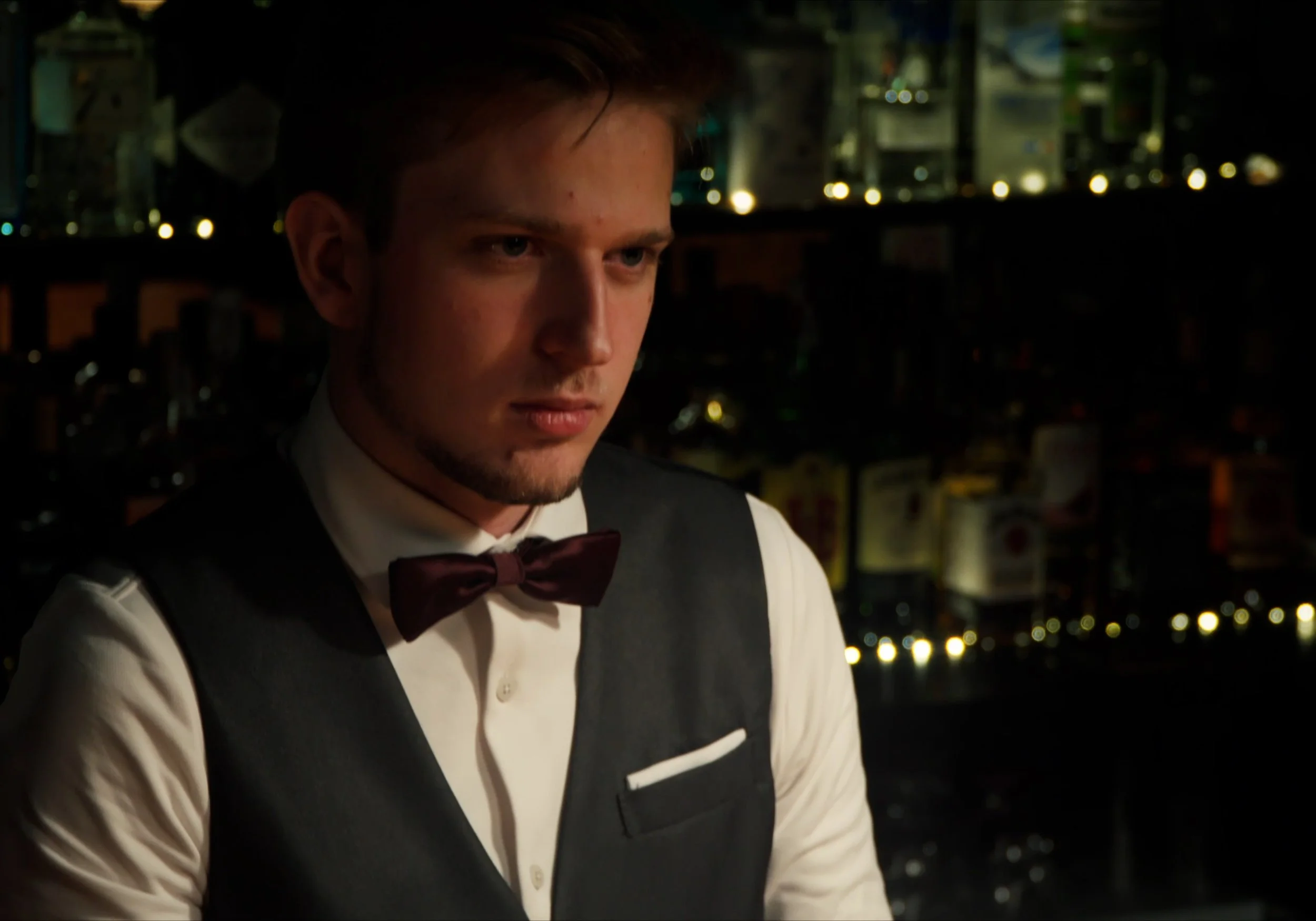 Un hombre joven con chaleco y pajarita en un bar con botellas al fondo.