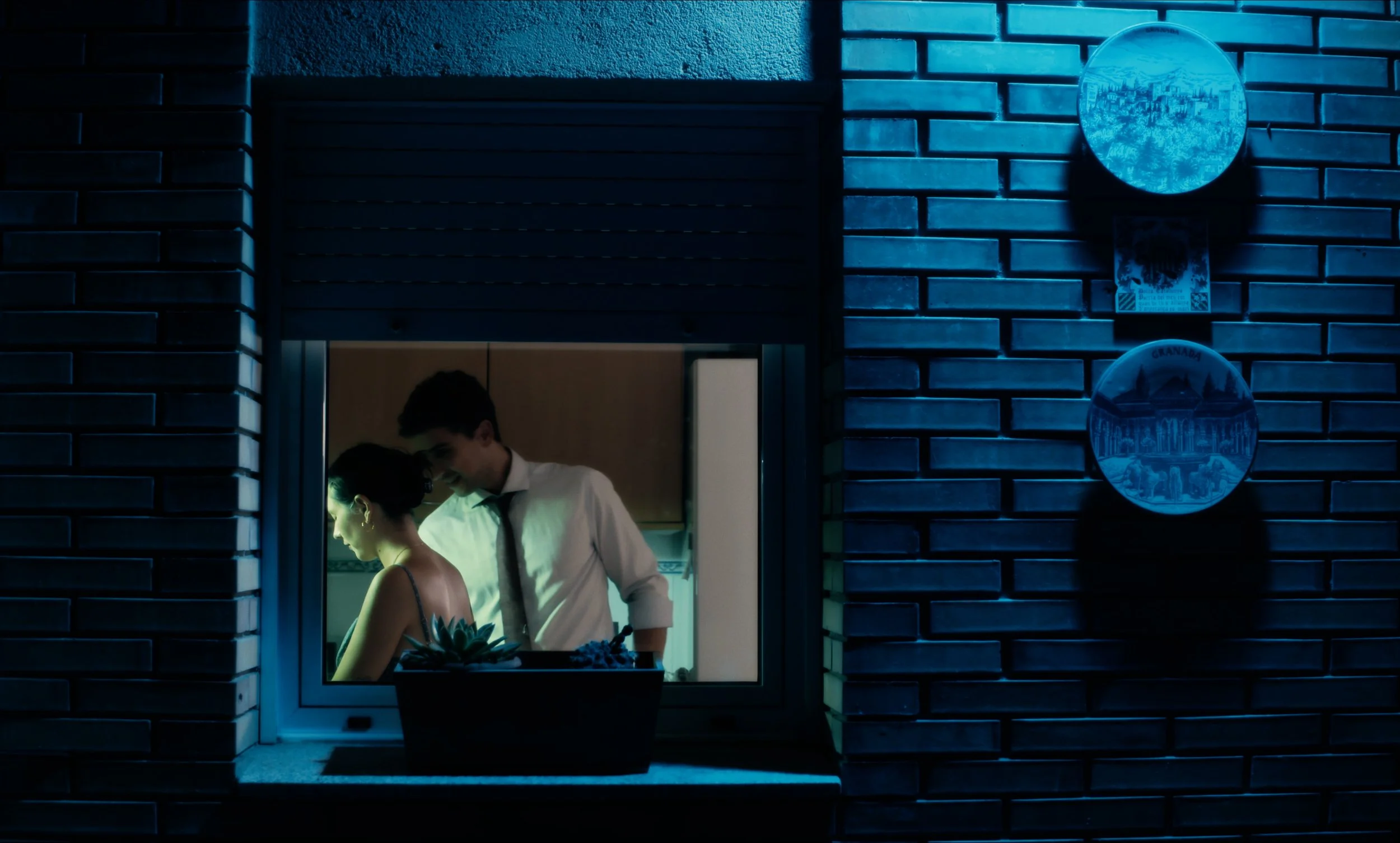 Dos personas en una cocina vistas a través de una ventana, con una pared de ladrillos y platos decorativos colgados afuera.