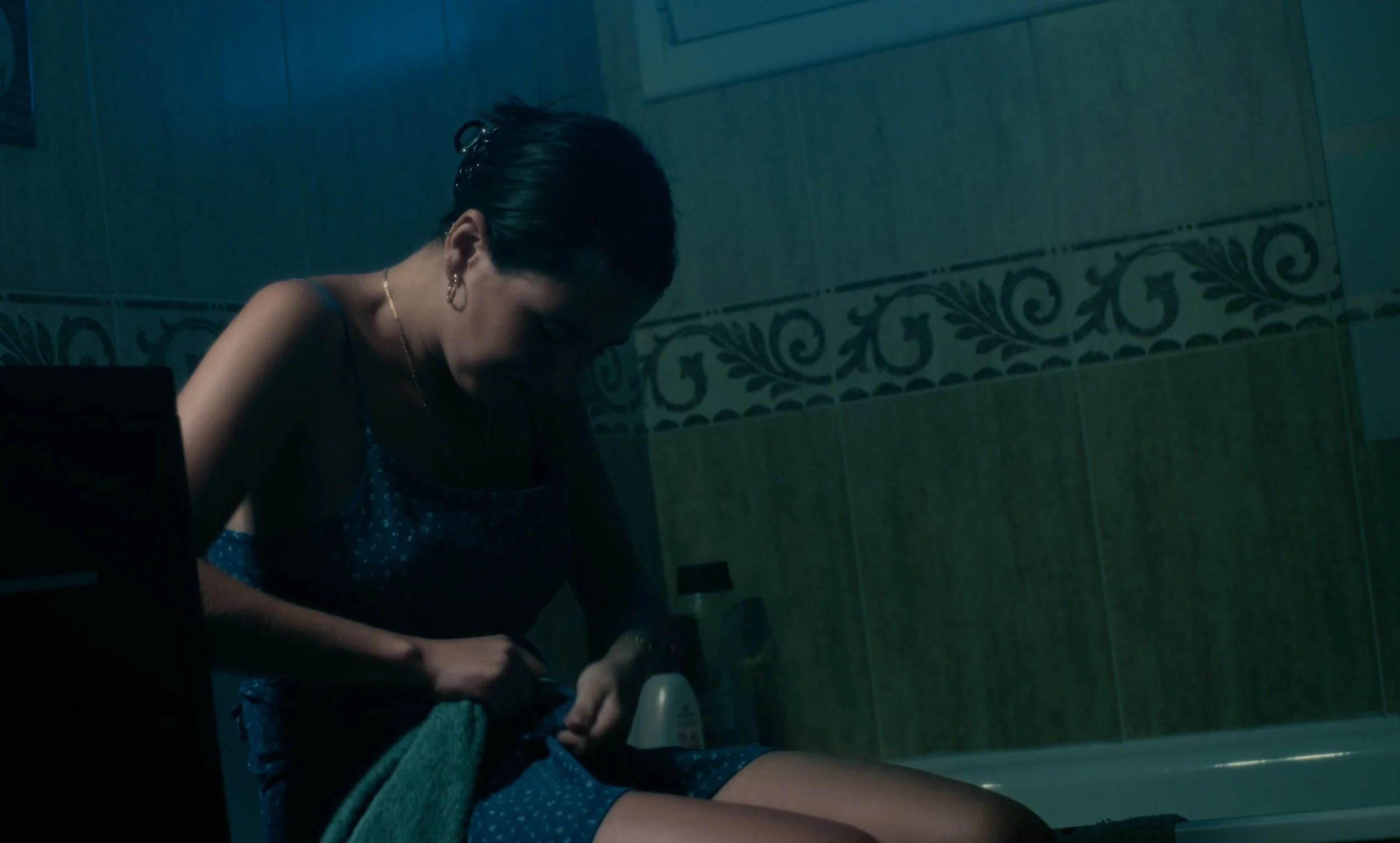 Mujer sentada en un baño con luz tenue, vistiendo un vestido de tirantes y sosteniendo una toalla verde.