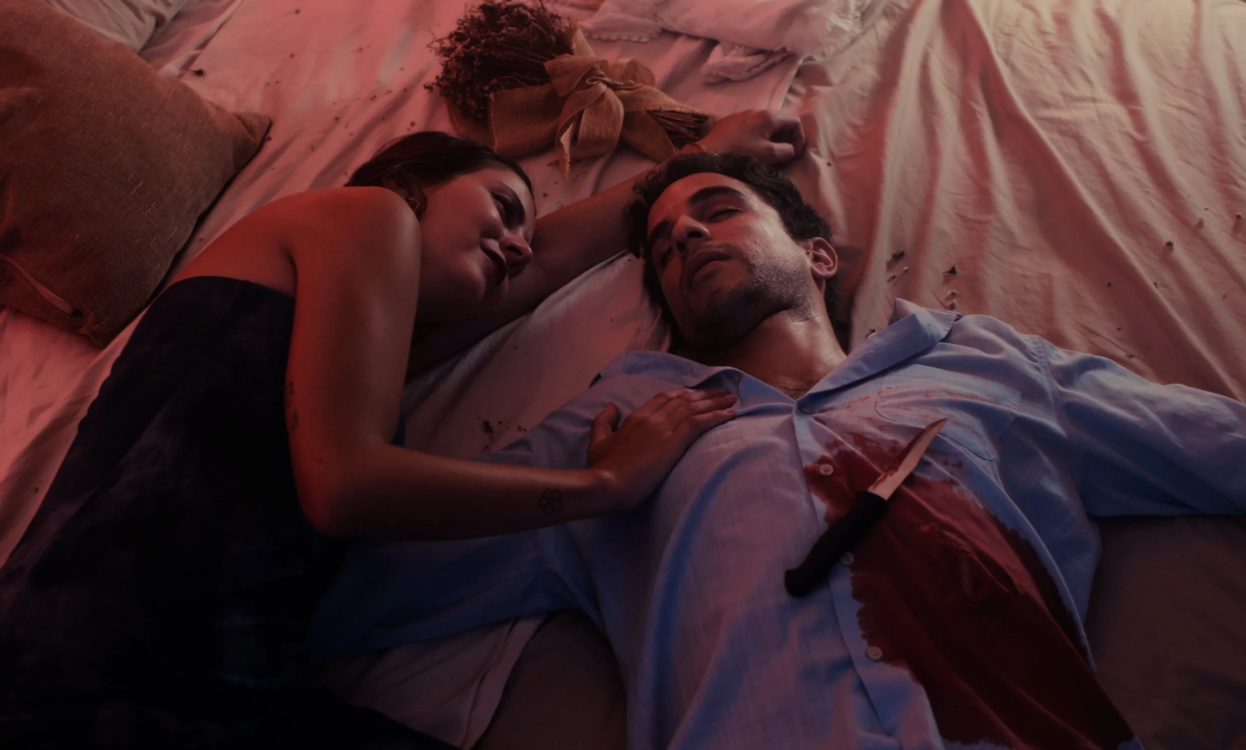 Una mujer y un hombre están acostados en una cama. El hombre tiene una herida con un cuchillo en el abdomen y su ropa está manchada de sangre. La escena tiene un tono dramático.