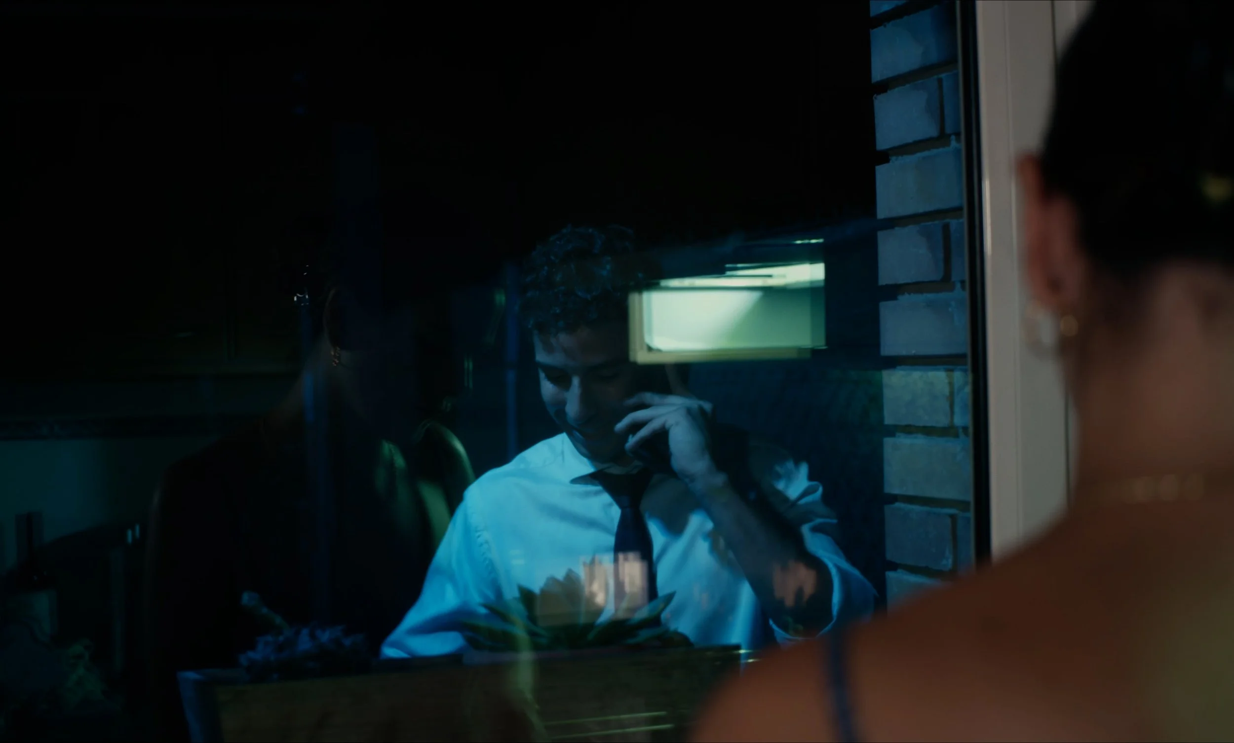 Un hombre en camisa blanca y corbata negra habla por teléfono en una habitación oscura. Un reflejo en la ventana muestra a una mujer de espaldas.