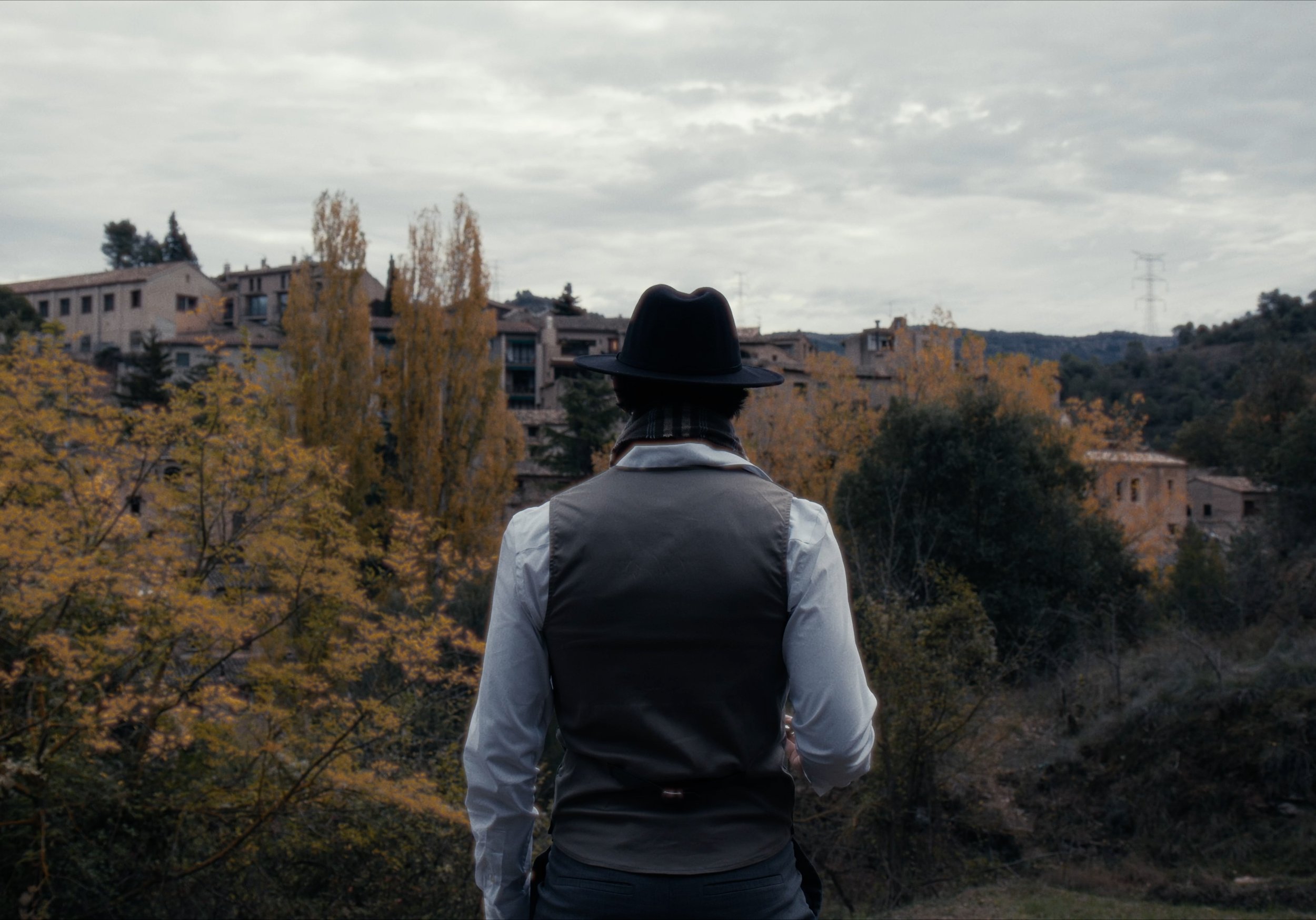 Persona de espaldas con sombrero, chaleco y chalina, mirando hacia un pueblo rodeado de árboles en otoño.