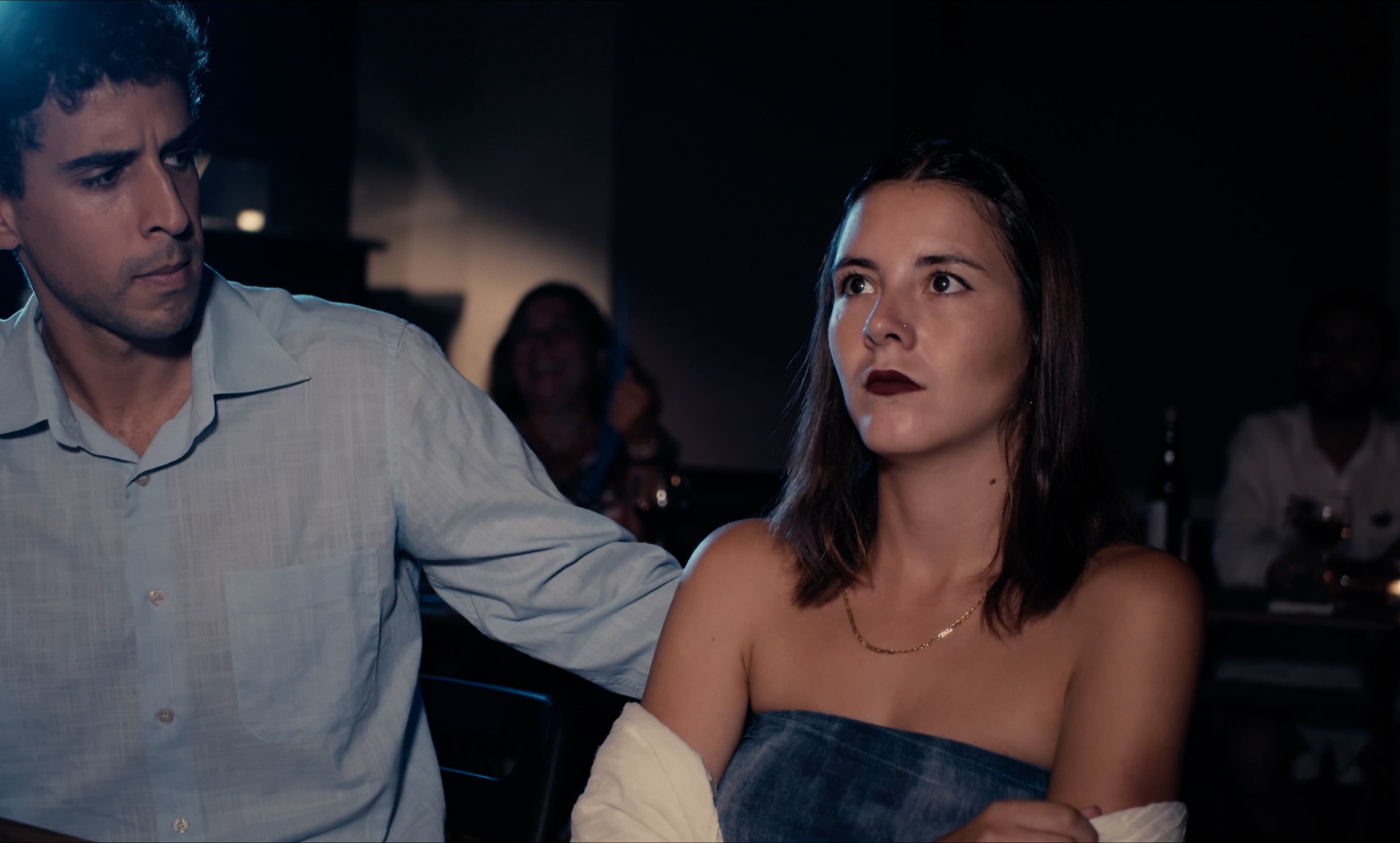 Un hombre y una mujer sentados en un ambiente oscuro. El hombre lleva una camisa clara y tiene expresión seria. La mujer lleva un top sin tirantes y tiene expresión pensativa. Se ven personas y botellas en el fondo.