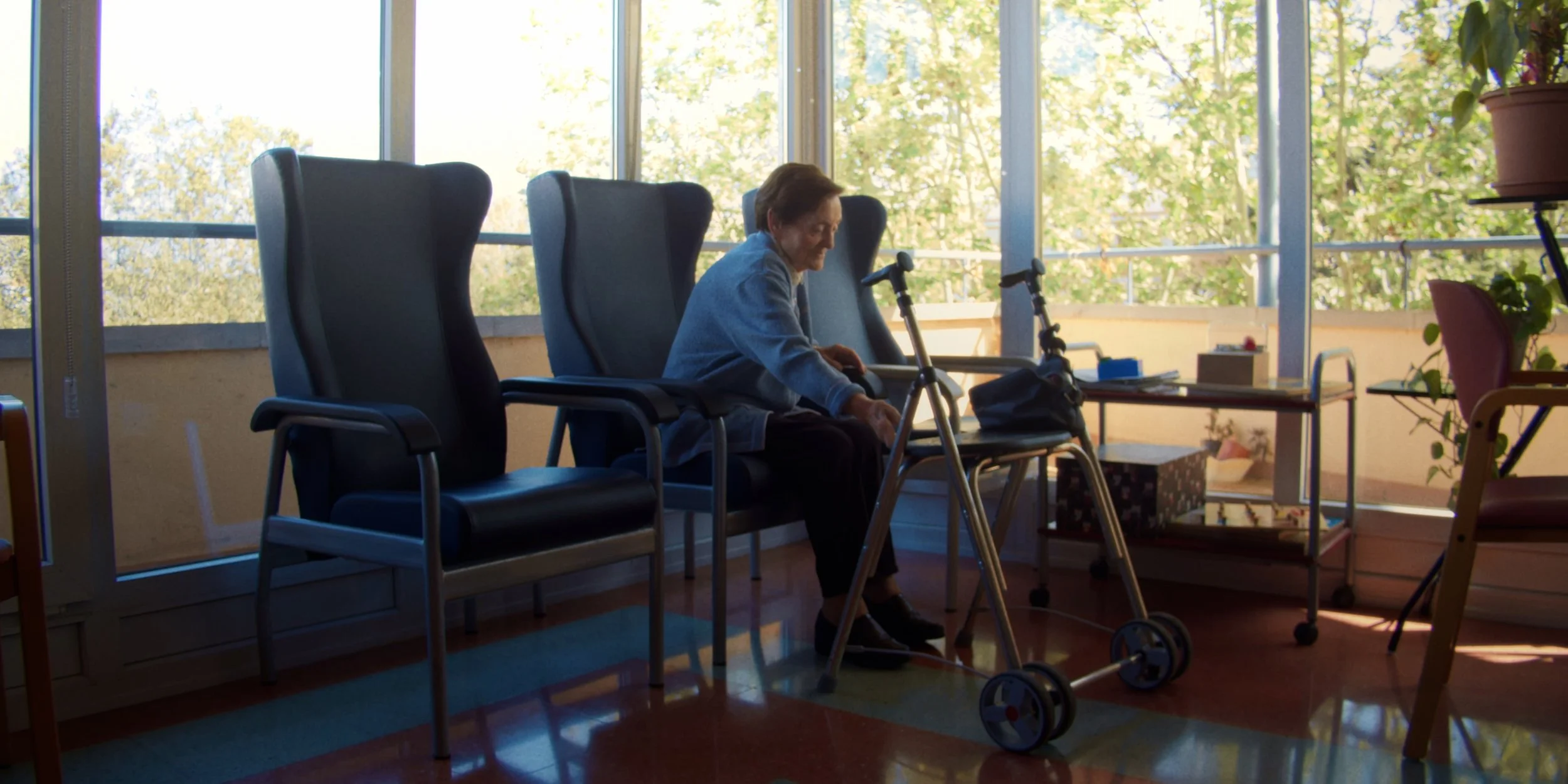 Una persona mayor sentada en una silla en un ambiente luminoso con ventanas grandes. Junto a ella, hay un andador y varias sillas adicionales. Al fondo, se observan plantas y una mesa con objetos. La escena parece ser en una sala de espera o un centro de atención para personas mayores.