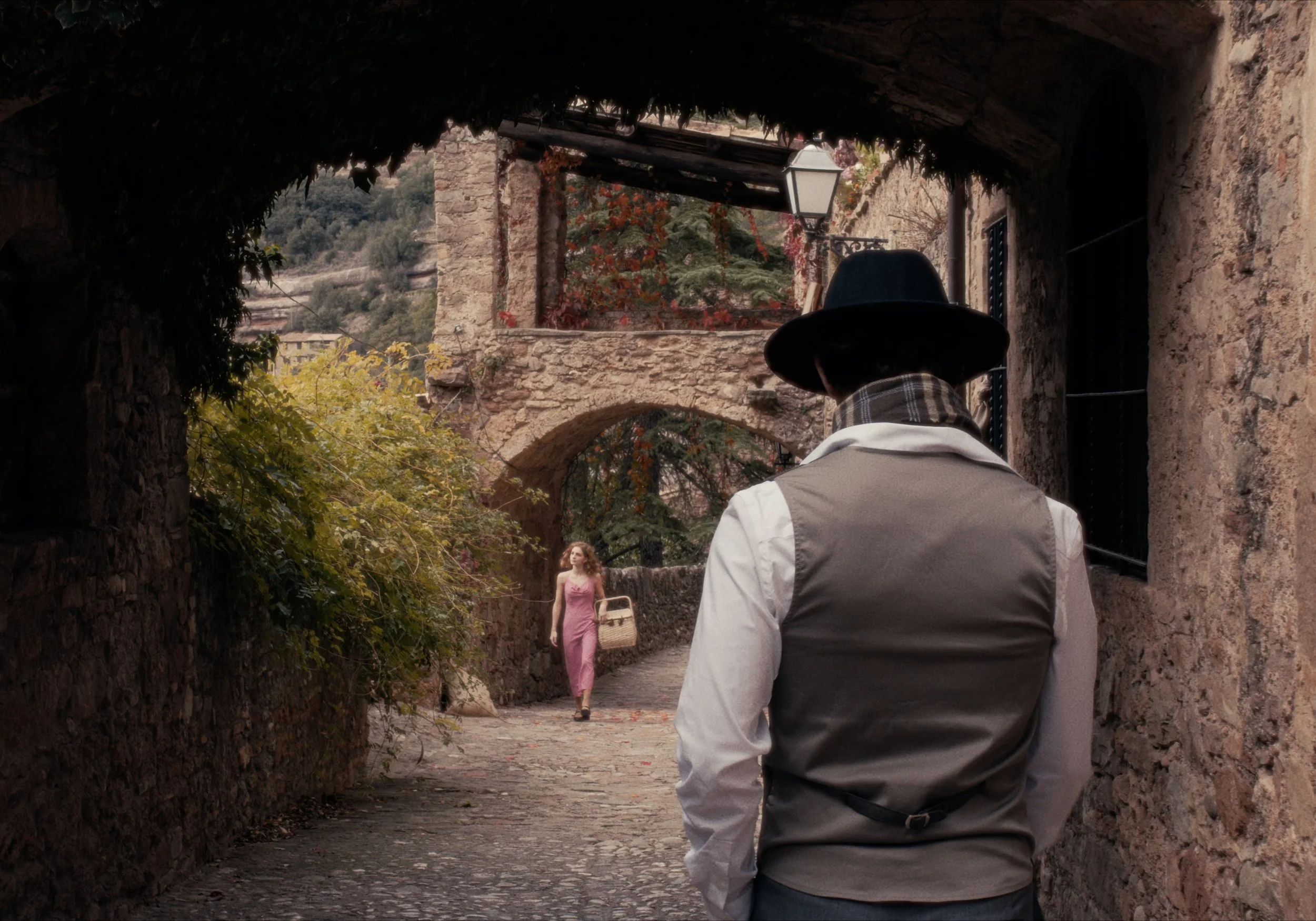 Persona con sombrero y bufanda observando a una mujer que camina por un callejón empedrado en un pueblo rústico.