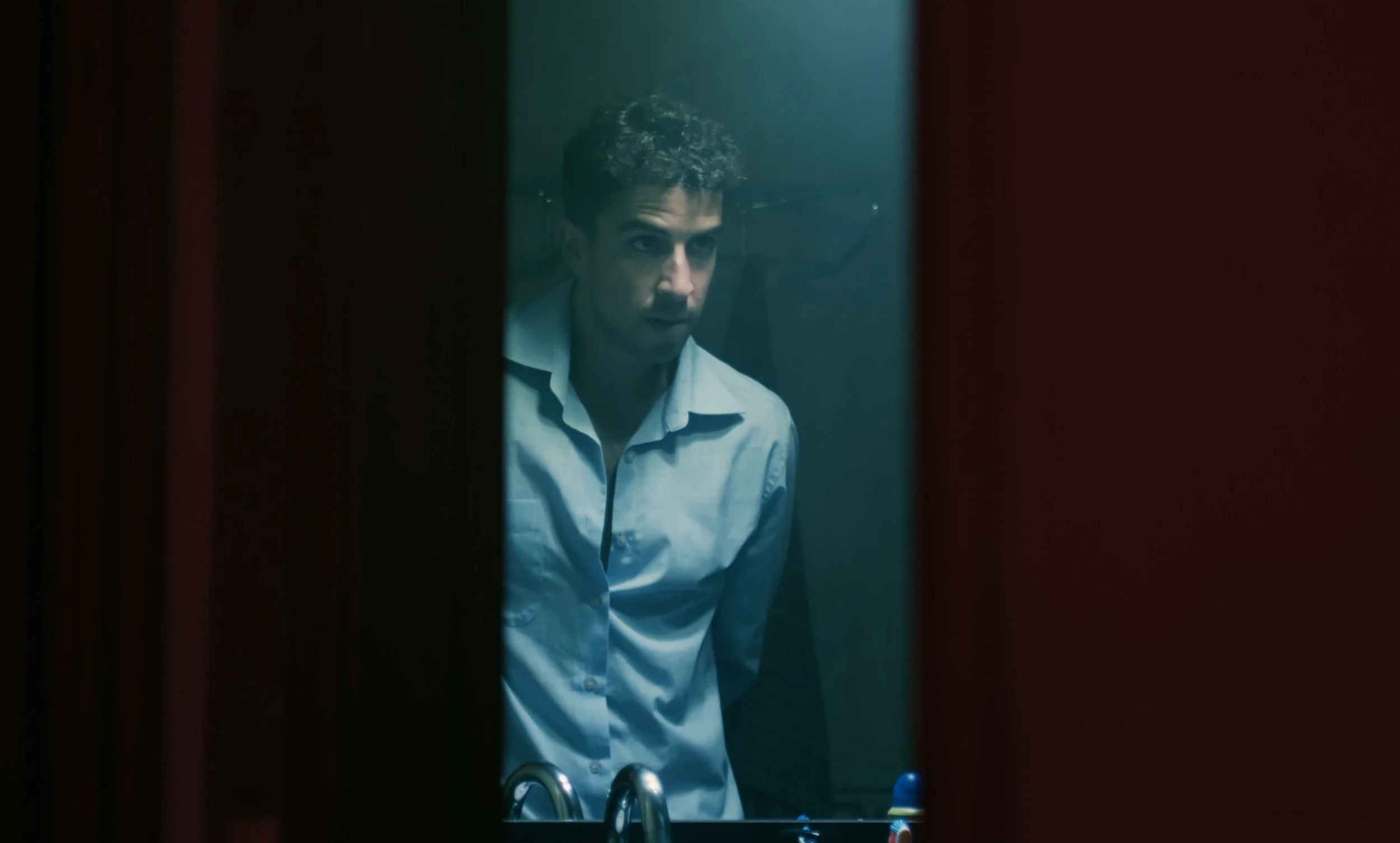 Hombre reflejado en el espejo del baño con camisa azul, iluminación tenue.