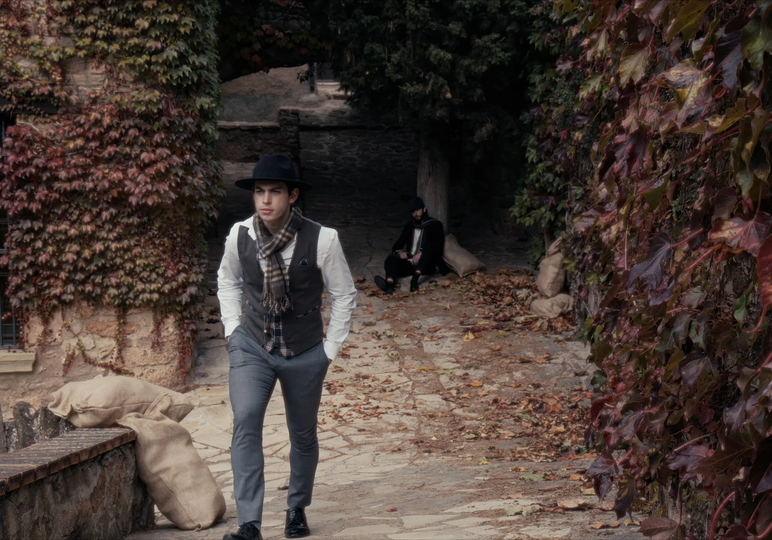 Hombre con sombrero y bufanda caminando por un camino empedrado, hojas caídas y paredes cubiertas de hiedra. Al fondo, otro hombre sentado.