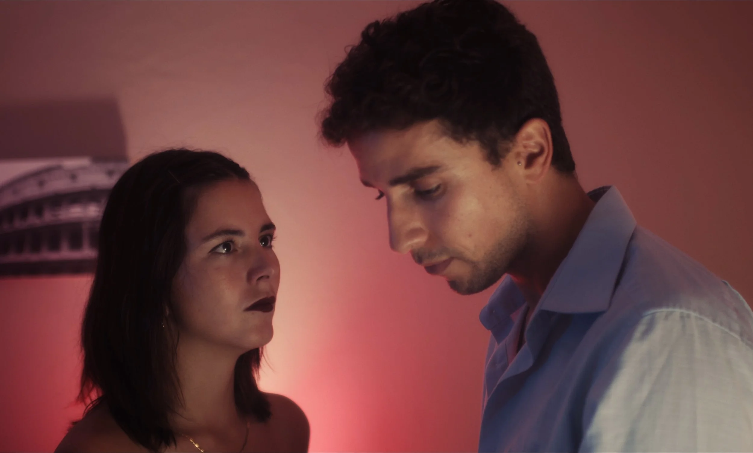 Una mujer y un hombre en una habitación con iluminación tenue, con la mujer mirando al hombre con una expresión seria.