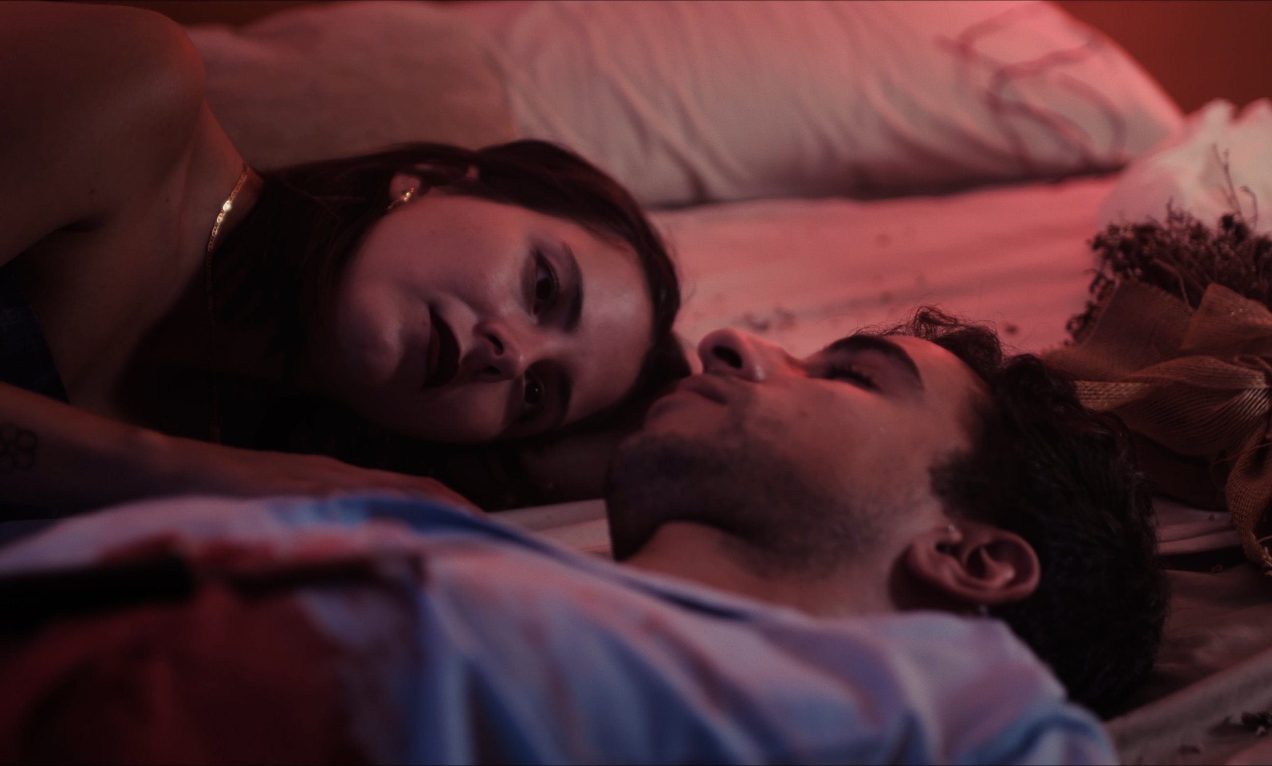 Dos personas acostadas en una cama, mirándose. El ambiente tiene iluminación tenue y colores cálidos.