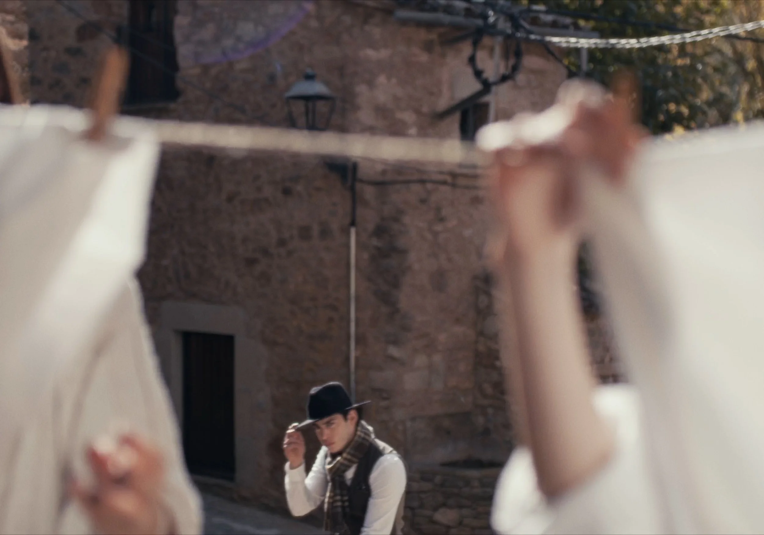 Hombre con sombrero negro y bufanda mirando a través de ropa colgada en una cuerda, con fondo de pared de piedra y farol.