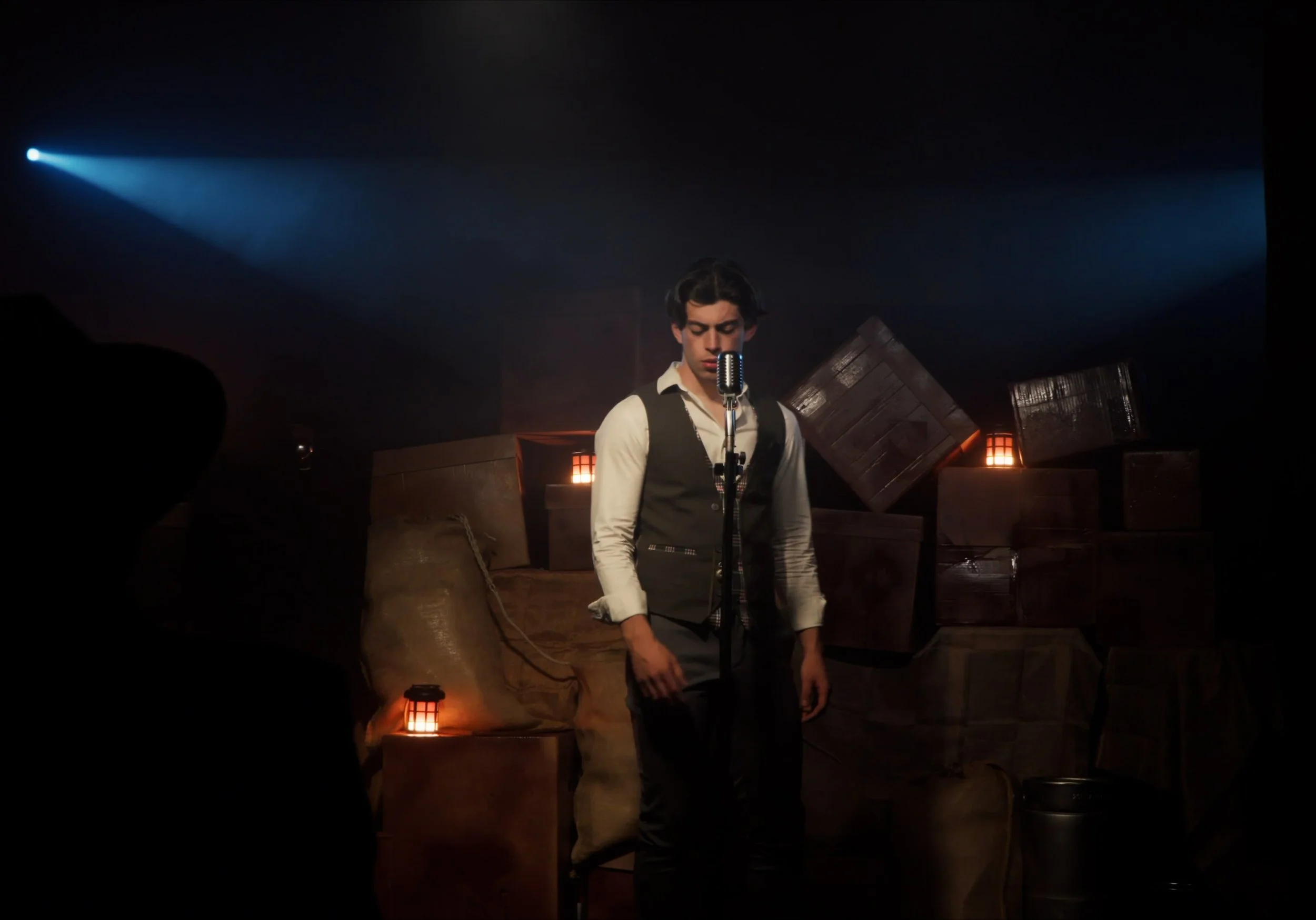 Un hombre joven con chaleco y camisa blanca está frente a un micrófono antiguo, con cajas y luces tenues de fondo en un escenario oscuro.
