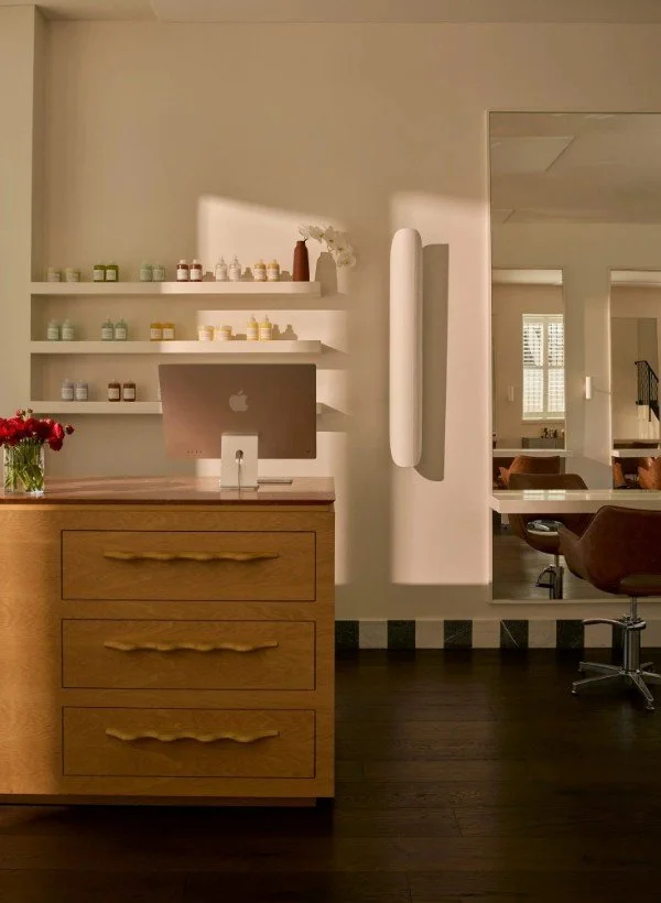 Interior of a modern salon with a wooden reception desk, computer, shelves with products, wall art, and salon chairs.