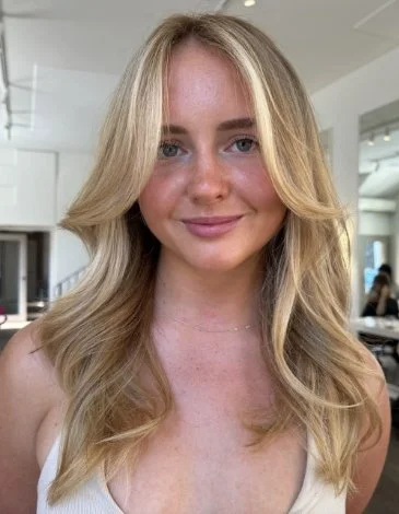Smiling woman with long wavy blonde hair in a light-colored top, standing indoors.