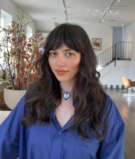 Person with long wavy hair wearing a blue shirt and a necklace, standing indoors near a plant.