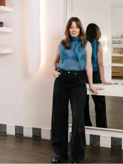 Woman standing in modern interior, wearing blue sleeveless top, black pants, leaning against a white counter. Shelves and mirror in the background.
