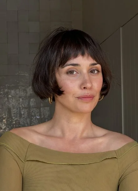 Woman with short dark hair, wearing an off-the-shoulder top, standing against a textured wall background.