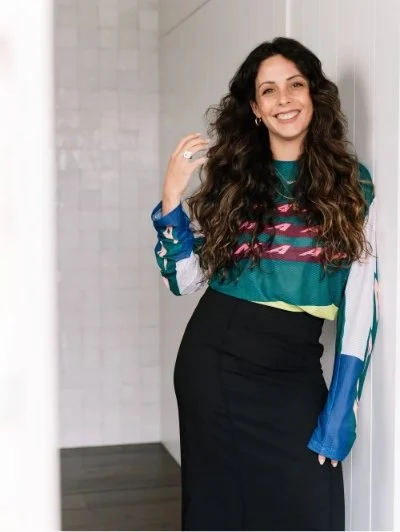 Smiling woman with long, curly hair wearing a colorful long-sleeve shirt and black skirt, standing in a bright indoor setting.