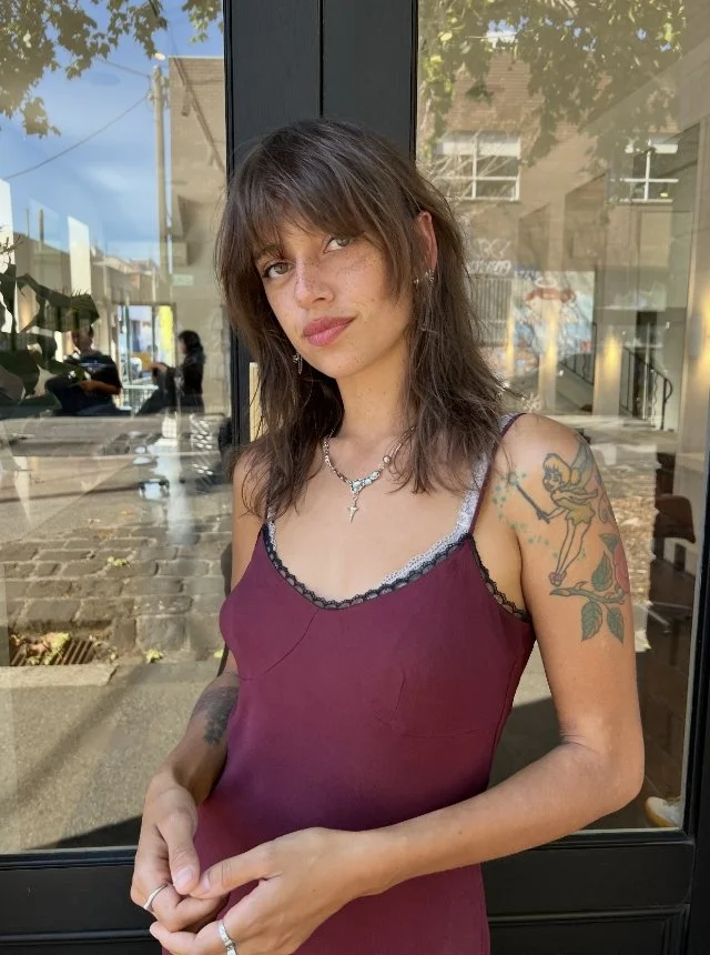 Portrait of a woman with brown hair and tattoos, wearing a burgundy camisole, standing outside near a glass door with reflections and urban background.