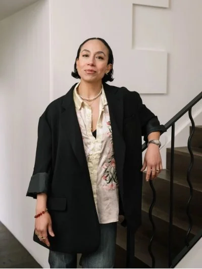 Person standing by a staircase wearing a black blazer, embroidered shirt, and jewelry.