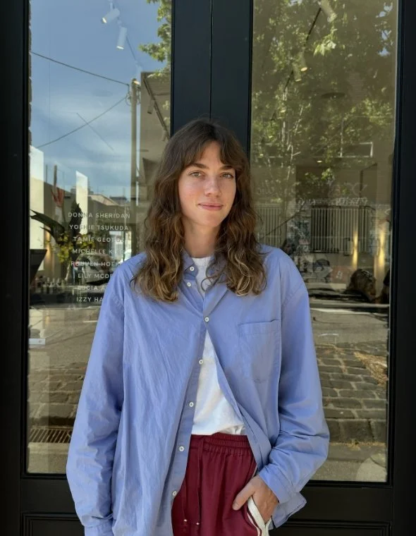 Person wearing a light blue shirt and red pants standing outside a glass building with reflections.
