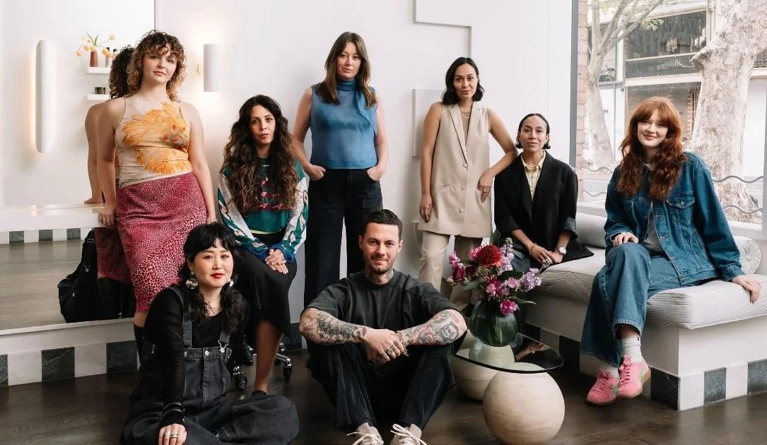 Group of eight people sitting and standing in a stylish room, showcasing a variety of fashion styles, with a vase of flowers in the foreground and large windows in the background.