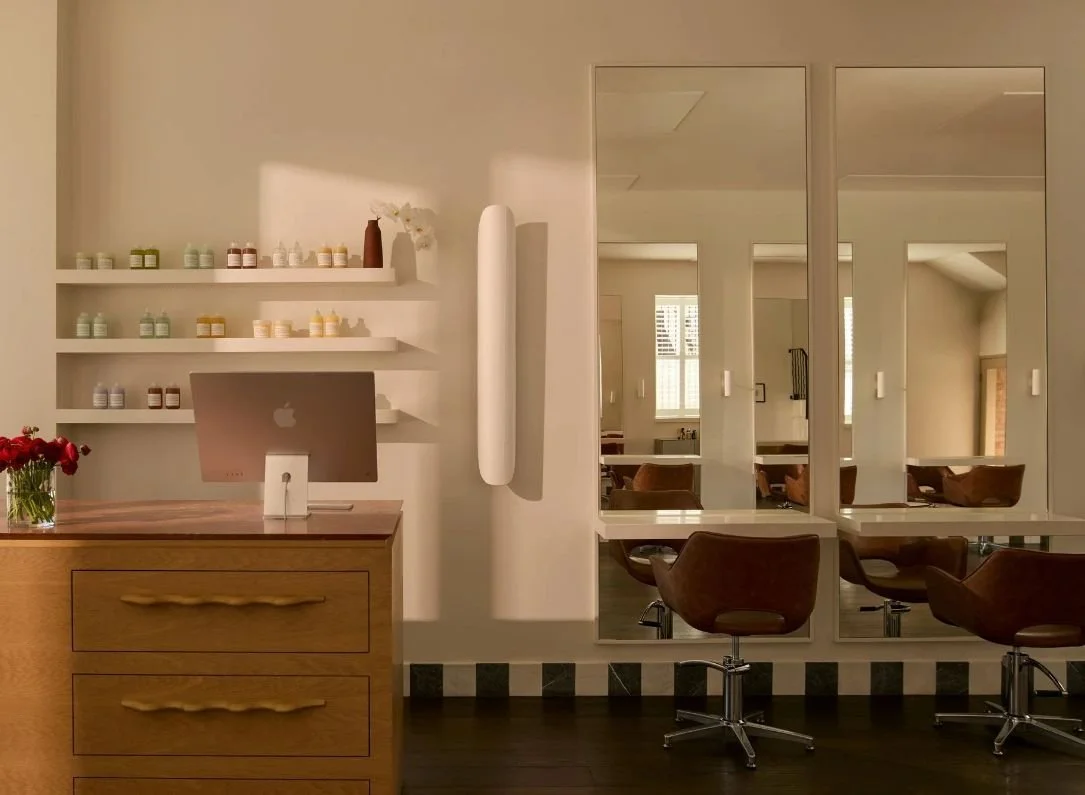 Modern salon interior with wooden furniture, mirrors, and styling chairs, featuring shelves with beauty products, a computer, and a vase with red flowers.