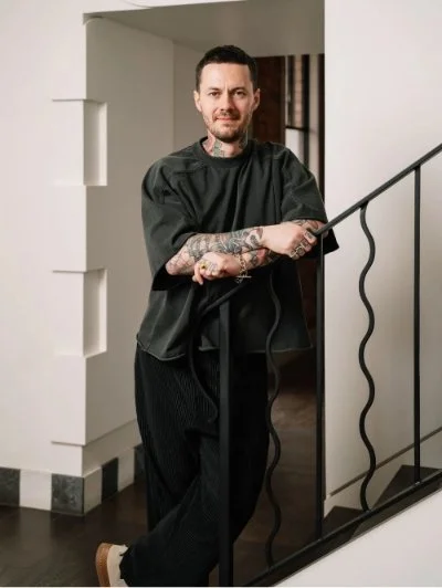 Person with tattoos standing on stairs, wearing a loose dark shirt and black pants.
