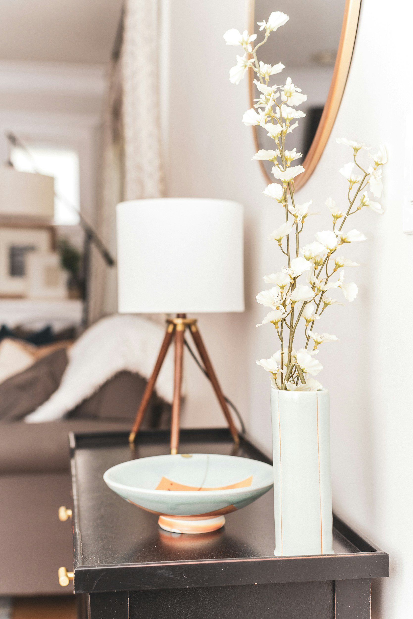 A tall white vase with white flowers on a black table, behind a table lamp with a white shade and wooden base, in a cozy living room.