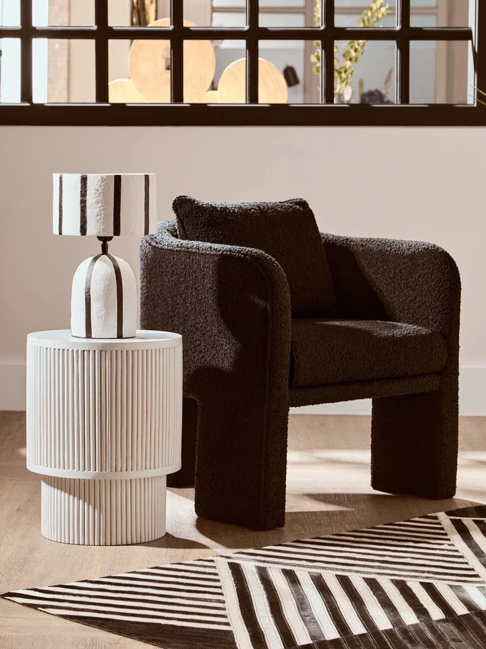 A black textured armchair with a matching pillow, a white ribbed round side table, and a black and white striped lamp in a modern interior.