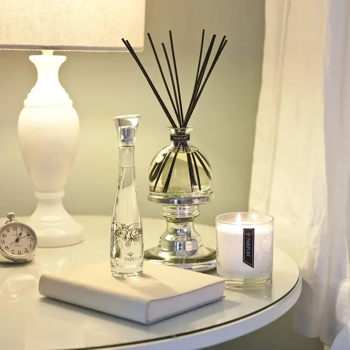 A bedside table with a white lamp, a round clock, a white book, a glass perfume bottle, a reed diffuser, and a lit candle.