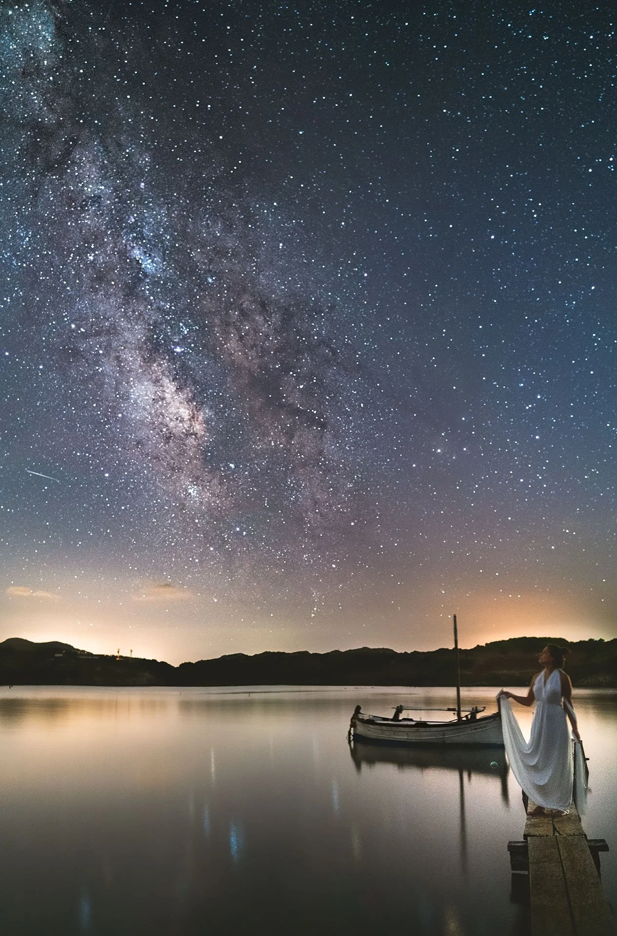 Mujer en vestido blanco sobre un muelle mirando el cielo estrellado y la Vía Láctea en una noche despejada, reflejándose en un lago tranquilo con una lancha a un lado.