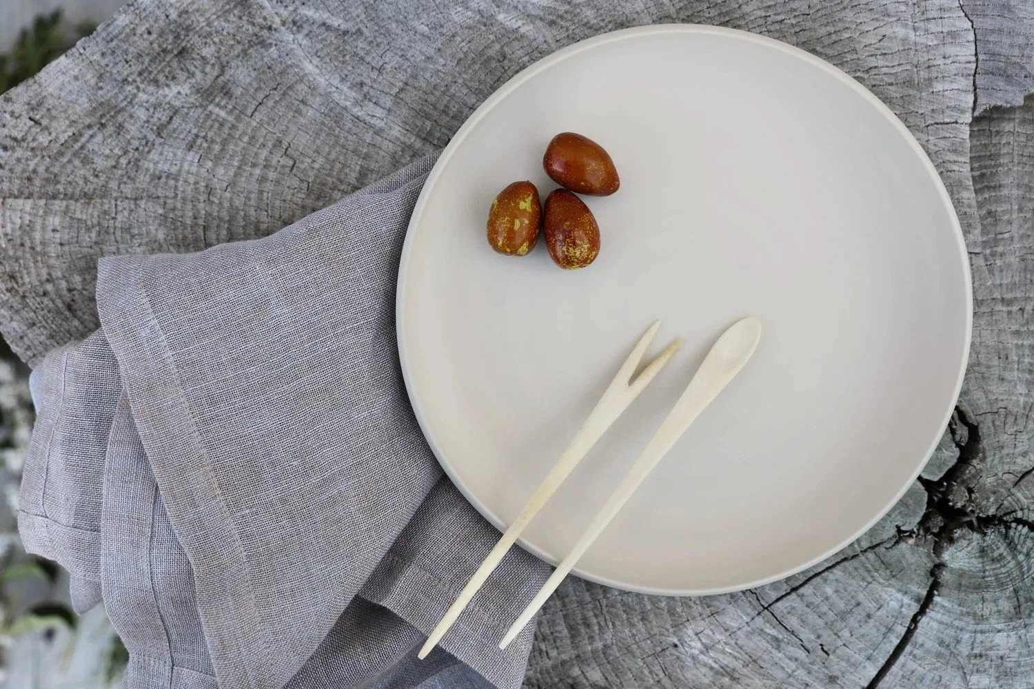 Stacked pastel-colored plates and bowls on a rustic wooden surface, with vintage silverware and a ceramic cup and saucer in the background, along with some green fruit on a leaf in the distance.