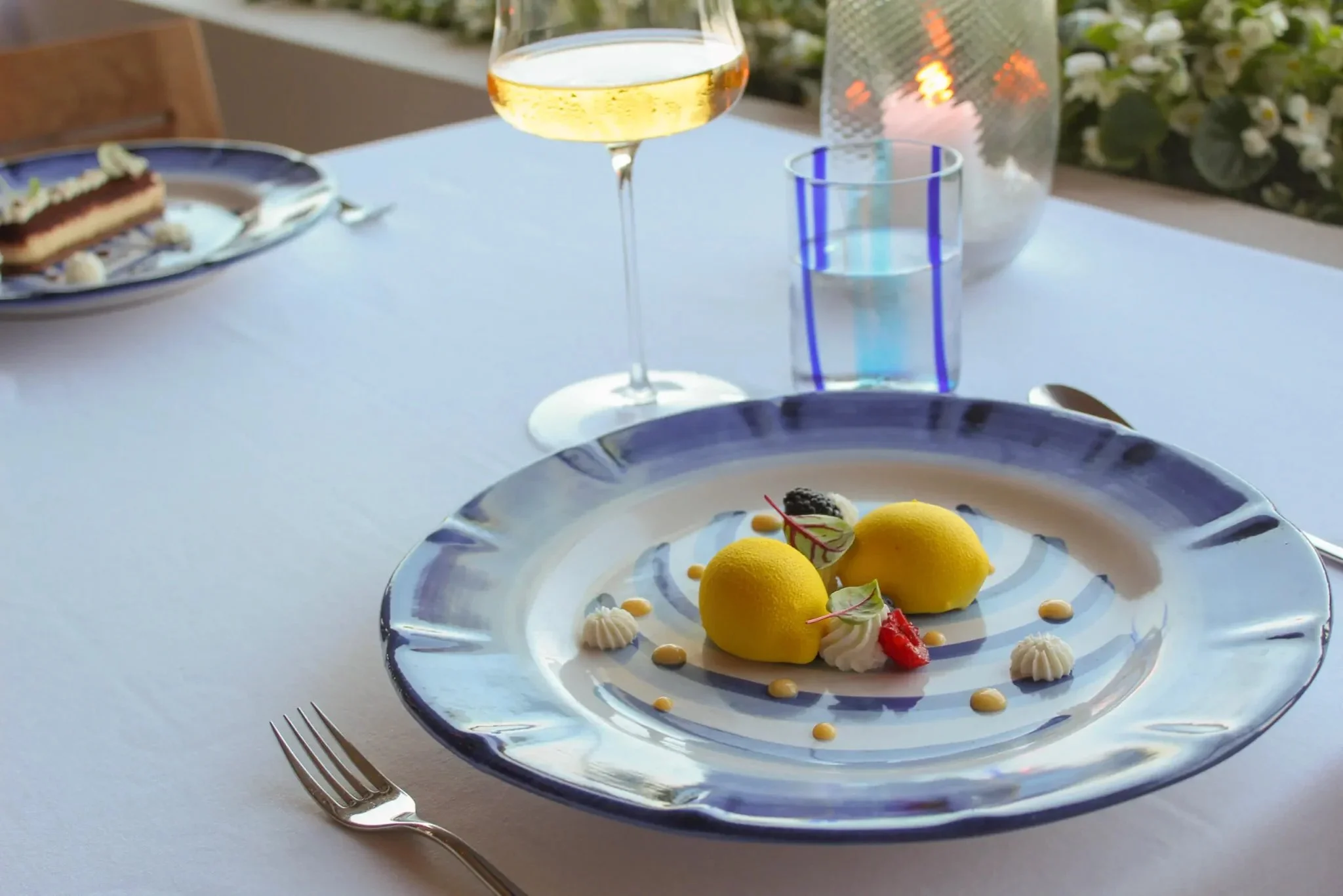 A plate with two yellow dessert scoops, garnished with berries, whipped cream, and small dollops of sauce on a white tablecloth. In the background, there is a glass of white wine, a water glass, and another similar plate.
