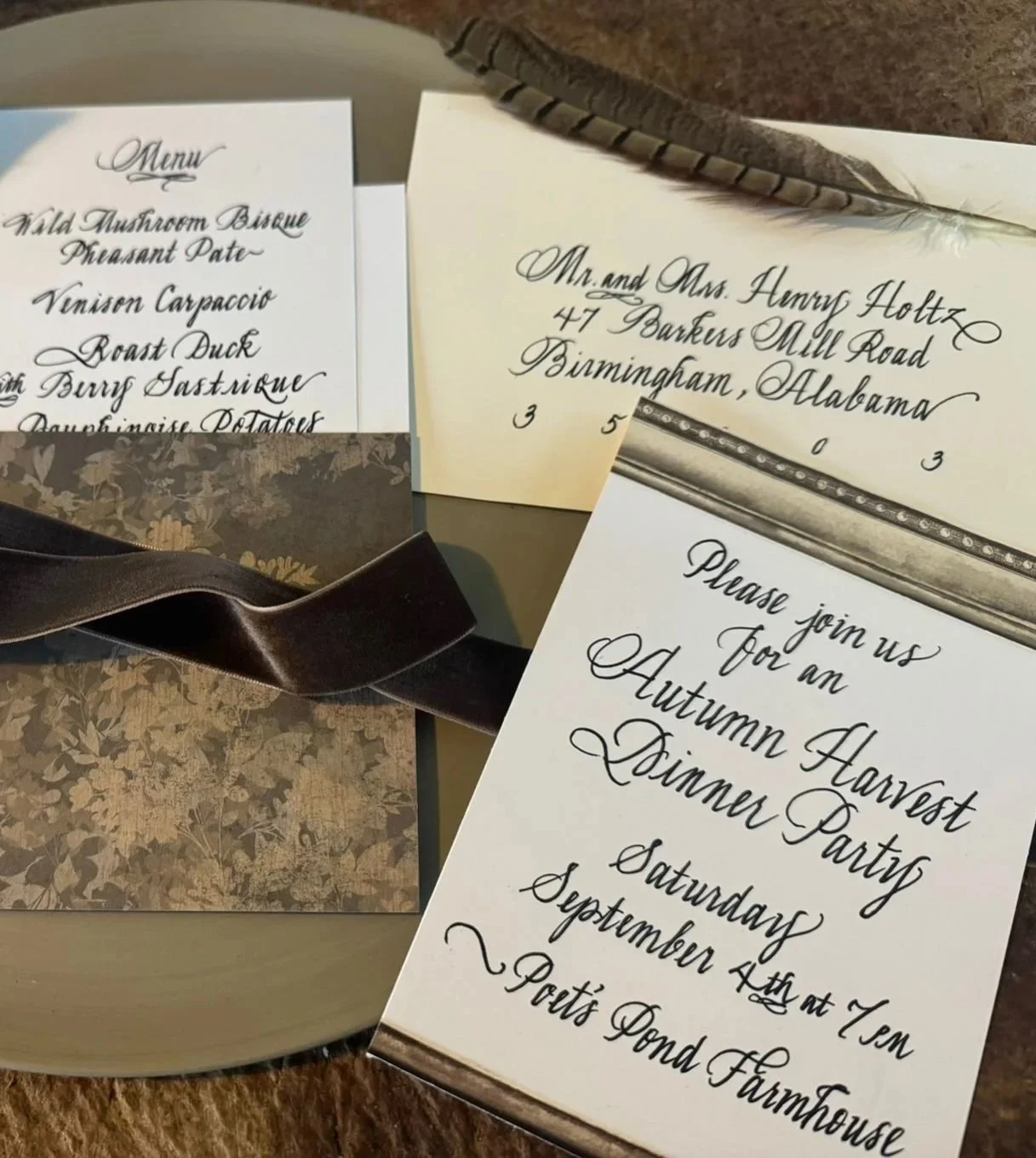 Invitation cards for an autumn harvest dinner party with handwritten script, one card with address, another with menu, and decorative elements including a feather and a ribbon, on a table.