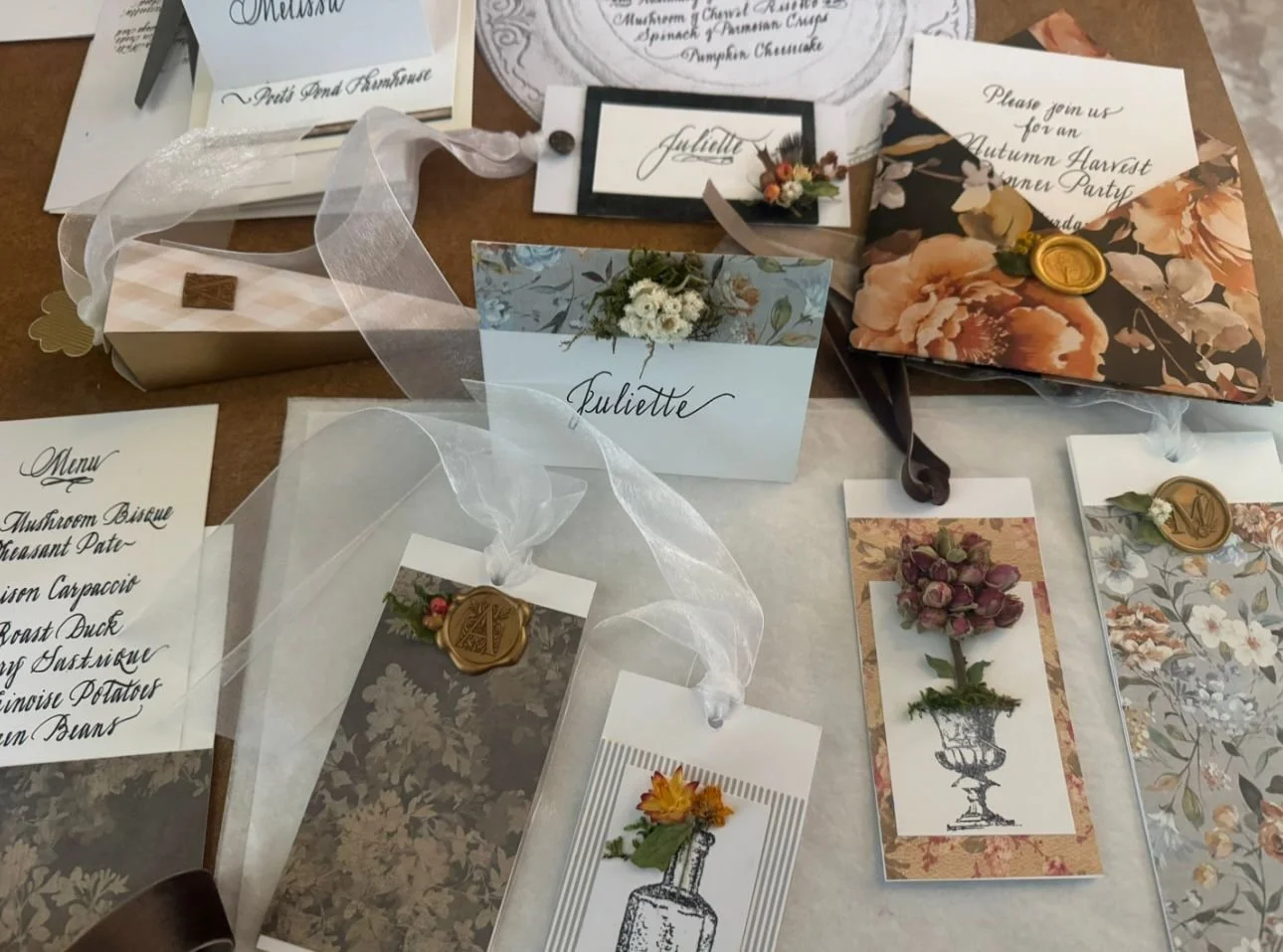 Decorative wedding or event stationery, including place cards, menus, and invites, featuring floral designs and gold wax seals, arranged on a table.