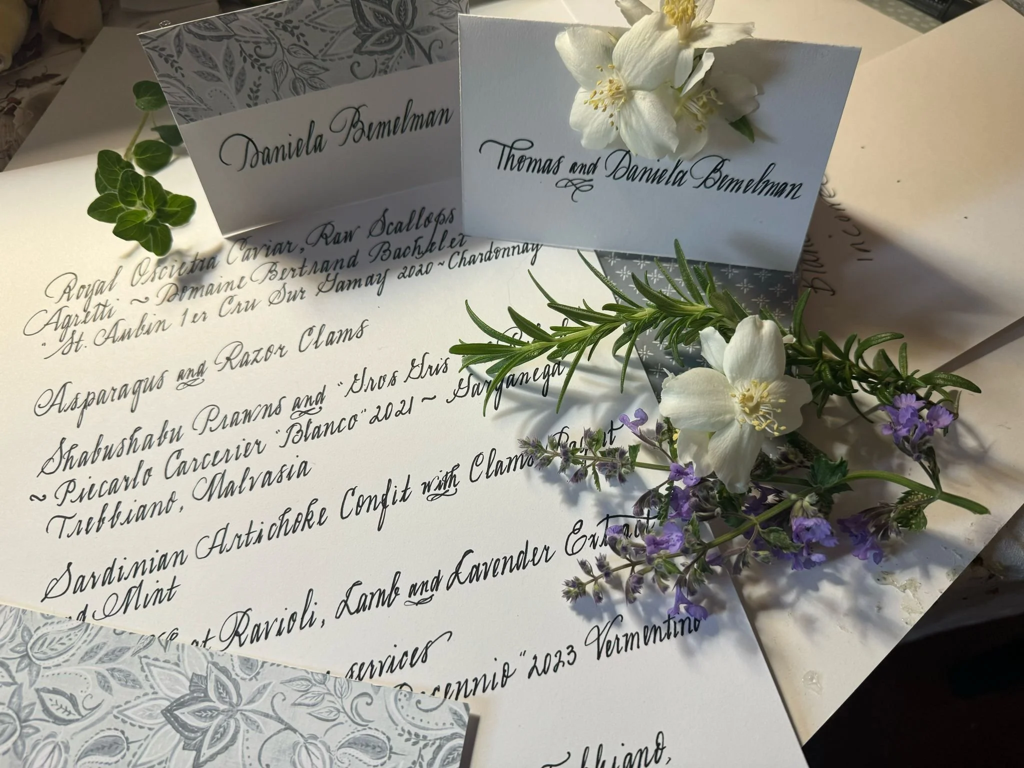 Wedding invitation with elegant black script text, decorated with white and purple flowers, including roses, lavender, and greenery.